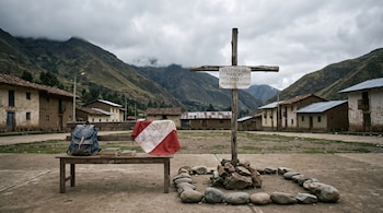 Una cruz de madera con cartel junto a piedras, y una banca con una mochila, libros y la bandera peruana frente a casas y montañas.