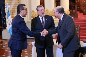 Tres hombres con traje y corbata, dos se dan la mano. Hay una bandera de Guatemala y una escalera con alfombra roja al fondo
