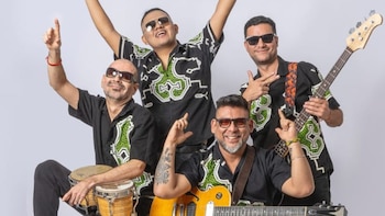 Cuatro hombres sonrientes con gafas de sol, camisas negras y verdes; uno con bongos, otro con guitarra eléctrica, otro con bajo eléctrico, posando