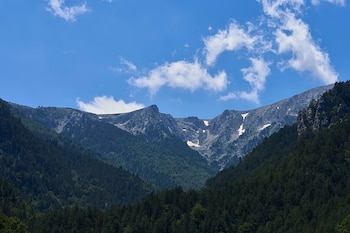 Montañas rocosas con picos, vegetación boscosa y parches de nieve, bajo un cielo azul con nubes blancas dispersas
