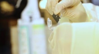 Primer plano de manos con guantes color crema manipulando un pequeño tubo transparente y un objeto negro. El fondo está desenfocado