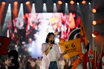 Keiko Fujimori de pie en un escenario iluminado, vistiendo un blazer rosa y jeans, hablando por un micrófono con un público borroso de fondo