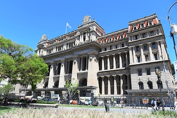 Edificio de piedra con columnas, techos de mansarda y bandera en la cima, protegido por vallas y andamios frente a una plaza con plantas