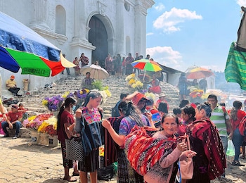Mercado concurrido en Chichicastenango. Numerosas personas en atuendos tradicionales, puestos con flores coloridas y sombrillas, frente a la fachada de una iglesia blanca