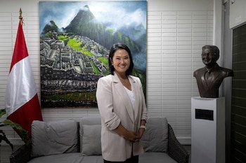 Keiko Fujimori, vestida con blazer claro, sonríe de pie en una sala junto a una bandera de Perú, un cuadro de Machu Picchu y un busto de su padre