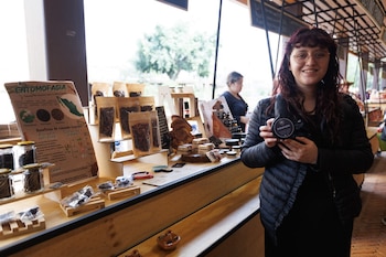 Una mujer con gafas sostiene un envase de pomada negra, de pie frente a un puesto de mercado con insectos comestibles y productos artesanales