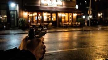 Unas manos en primer plano sostienen una pistola que apunta hacia una calle y un restaurante iluminado durante la noche.