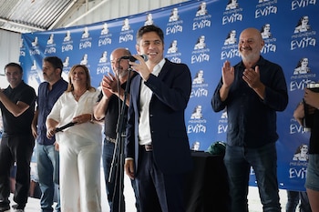Un hombre sonriente habla en un micrófono en el centro, rodeado de otras personas que aplauden. Detrás, un telón azul con el logo "Movimiento Evita"