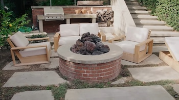 Patio exterior con fogata de ladrillo y piedras volcánicas, tres club chairs de madera clara y cojines blancos, un sofá y parrilla empotrada