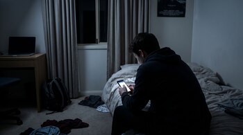 Persona de espaldas sentada en cama oscura mira teléfono móvil; se ven un escritorio con laptop, una mochila y ropa en el suelo.