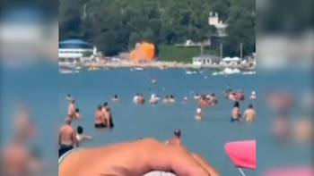 Una playa con muchos bañistas en el mar, una explosión naranja en la arena, edificios y árboles de fondo, y una mano con un objeto rosa en primer plano