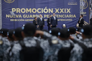 Autoridades en traje de vestir sobre un escenario frente a un letrero azul y agentes uniformados de espaldas en primer plano