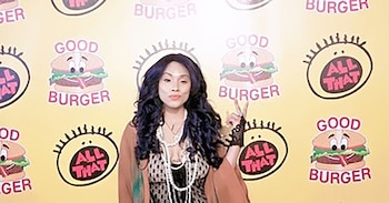 Mujer con cabello oscuro y rizado, collares de perlas, blusa estampada y chaqueta marrón posa ante fondo amarillo con logos de hamburguesas y caras sonrientes