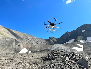 Helicóptero multirotor blanco y negro con hélices rojas y negras en vuelo sobre un valle montañoso rocoso con nieve, bajo un cielo azul despejado