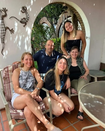 Un hombre, una mujer y tres jóvenes sonríen en un patio con sillas de terraza, plantas y un arco de ladrillo al fondo