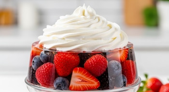 Un postre en vaso transparente con gelatina, fresas enteras y en rodajas, arándanos, frambuesas, moras y crema batida encima.