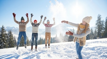 Cuatro niños y una mujer en ropa de invierno saltan y lanzan nieve al aire en un paisaje nevado, con montañas y pinos bajo un cielo azul brillante.