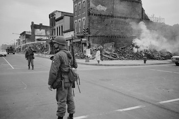Vista en blanco y negro de soldados en una calle. Un militar armado de espaldas, otro rociando con manguera, y un edificio en ruinas con humo a la derecha