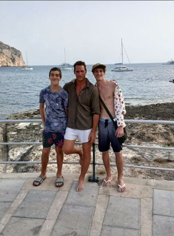 Tres hombres de pie sobre un piso de baldosas junto a una barandilla metálica, con el mar, barcos y una montaña al fondo