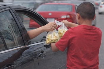 Un niño de espaldas con camiseta roja ofrece dos bolsas de fruta picada a un brazo que se asoma por la ventanilla de un coche oscuro. Vehículos al fondo.
