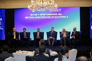 Seis hombres sentados en un escenario frente a una pantalla con texto. Uno de ellos habla por micrófono. Hay un público sentado en mesas delante del escenario