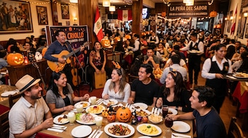 Personas cenando comida peruana en una mesa decorada con calabazas de Halloween mientras dos músicos tocan la guitarra y el cajón en un local.