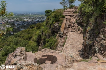 Escalinatas y estructuras de piedra talladas en la roca de un sitio arqueológico con vegetación, una ciudad distante y cielo azul. Logo de INAH