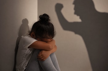 Una niña sentada en el suelo de un rincón con la cabeza agachada frente a la sombra proyectada de un adulto con el puño levantado.