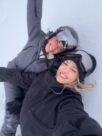 Dos mujeres con indumentaria de invierno y antiparras de esquí sonríen recostadas sobre la nieve