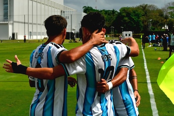 Tres futbolistas con camisetas a rayas celestes y blancas se abrazan de espaldas en un campo de césped verde, con edificios blancos y vegetación de fondo
