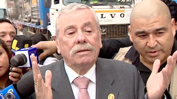 Un hombre canoso con bigote usa traje y corbata mientras habla a cuatro micrófonos. Está rodeado por personas, incluyendo un hombre calvo. Un camión Volvo en el fondo.