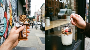 Mano sostiene una barrita de proteína con envoltorio de aluminio en un fondo urbano; otra mano sostiene una cuchara dentro de un vaso de yogur con granola y bayas.