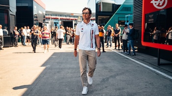 Hombre con polo blanco, pantalones caqui y gafas de sol camina en una pista. Hay personas y camiones de equipos en el fondo.