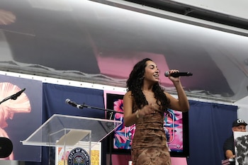 Una mujer joven con vestido estampado sostiene un micrófono mientras canta junto a un atril transparente con el sello de Miami