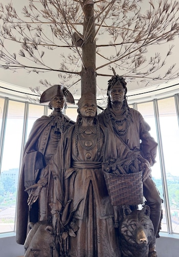 Escultura de bronce con hombre en sombrero tricorne, mujer y hombre indígena con cesta de maíz. Tronco y ramas de árbol metálico sobre ellos. Ventanas al fondo