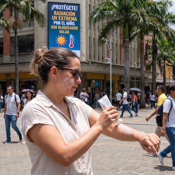 Una mujer aplica crema solar en su brazo en una plaza peatonal frente a un cartel publicitario sobre radiación ultravioleta.