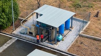 Vista aérea de una estructura con techo metálico, tanques azules, tuberías y dos personas de pie junto a un depósito de agua