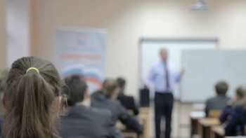 Vista trasera de estudiantes sentados en pupitres, escuchando a un profesor de pie frente a una pizarra en un aula