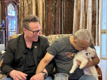 Rafi, con un pin de corazón arcoíris en la solapa, sonríe mientras Ashraf besa a un perro pomerania blanco, sentados en un sofá estampado (Fuente Latina)