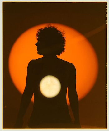 Silueta oscura de una persona con cabello rizado de espaldas, mirando a la izquierda, contra un gran círculo de luz naranja y uno pequeño blanco en su espalda