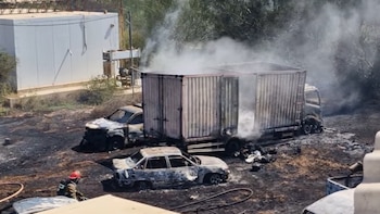 Vehículos calcinados, incluyendo un camión humeante y dos automóviles, en un terreno carbonizado. Un bombero con casco rojo trabaja en el área de restos quemados