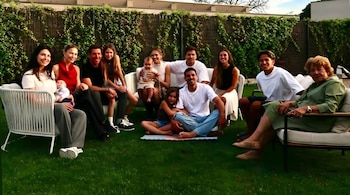 Vista amplia de una familia multigeneracional posando en un jardín con césped verde y un seto alto; algunos sentados en sofás blancos y otros en el suelo.