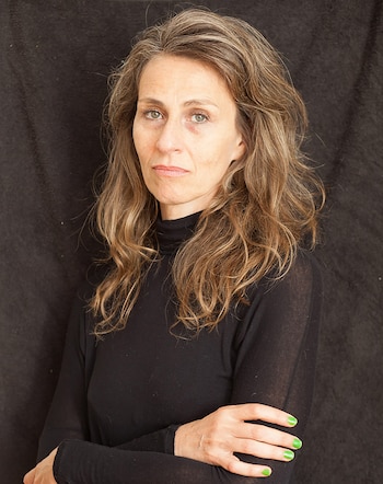 Primer plano de una mujer caucásica de mediana edad, con cabello castaño claro y canas, ojos verdes, y un suéter de cuello alto negro, con los brazos cruzados y las uñas pintadas de verde neón