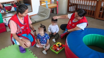 Dos mujeres con chalecos rojos de cuidadoras atienden a dos niños pequeños sentados en el suelo junto a una cesta de juguetes