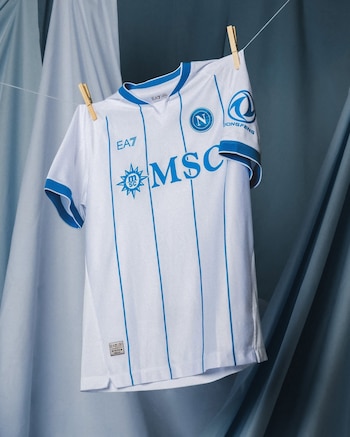 Camiseta de fútbol blanca y azul con rayas verticales, escudo del Napoli y patrocinadores EA7, MSC, DONGFENG, colgada de una soga con broches de madera