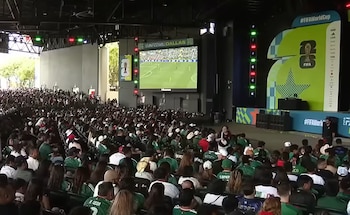 Vista aérea de una multitud sentada en un fan festival de fútbol con una gran pantalla mostrando un partido y un escenario con logotipos de FIFA World Cup