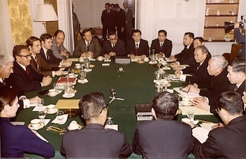 Un grupo de personas vestidas con traje se sienta alrededor de una mesa redonda cubierta con un mantel verde en una sala