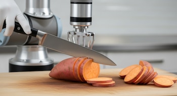 Primer plano de un brazo robótico metálico con un guante blanco que sostiene un cuchillo de chef cortando una batata en rodajas finas sobre una tabla de madera.