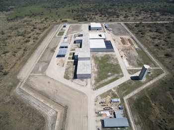 Vista aérea de un extenso complejo industrial con múltiples edificios rectangulares, carreteras de acceso, áreas de tierra arada y una torre alta, rodeado de vegetación