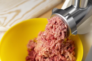 Una picadora de carne procesa carne cruda, que sale finamente molida y cae en un tazón amarillo sobre una superficie de madera clara